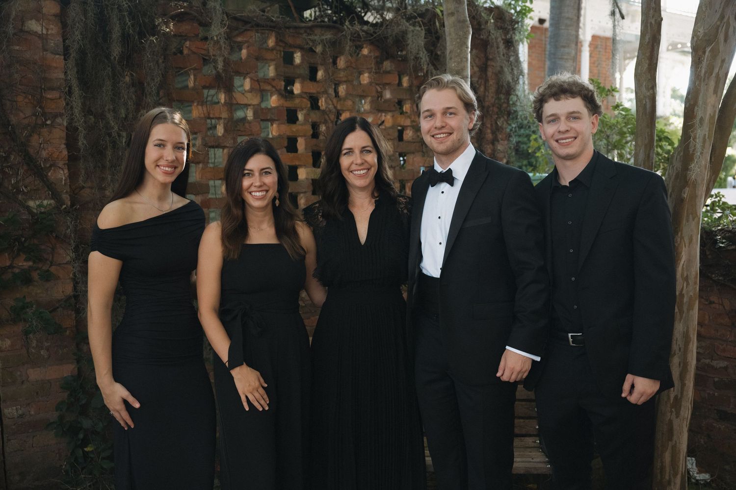 Five elegantly dressed people in black formal attire smiling together outdoors at a formal event.