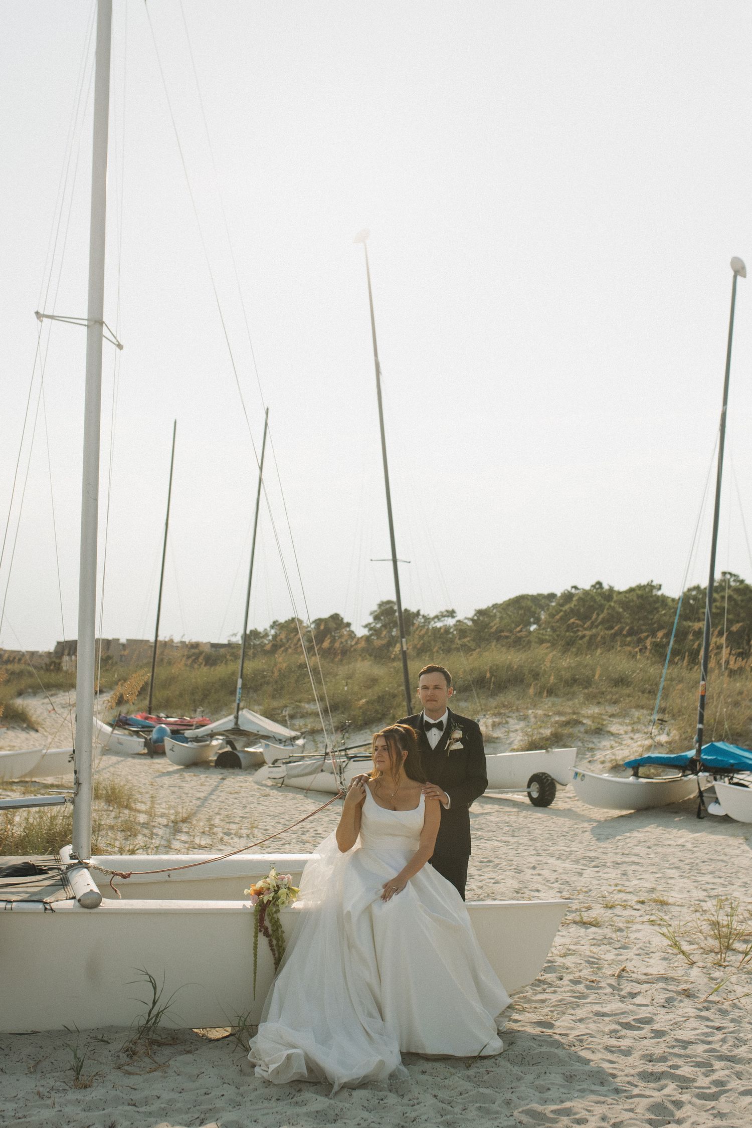 Bride in white gown sits beside groom in tuxedo on sandy beach with sailboats and dunes in background.