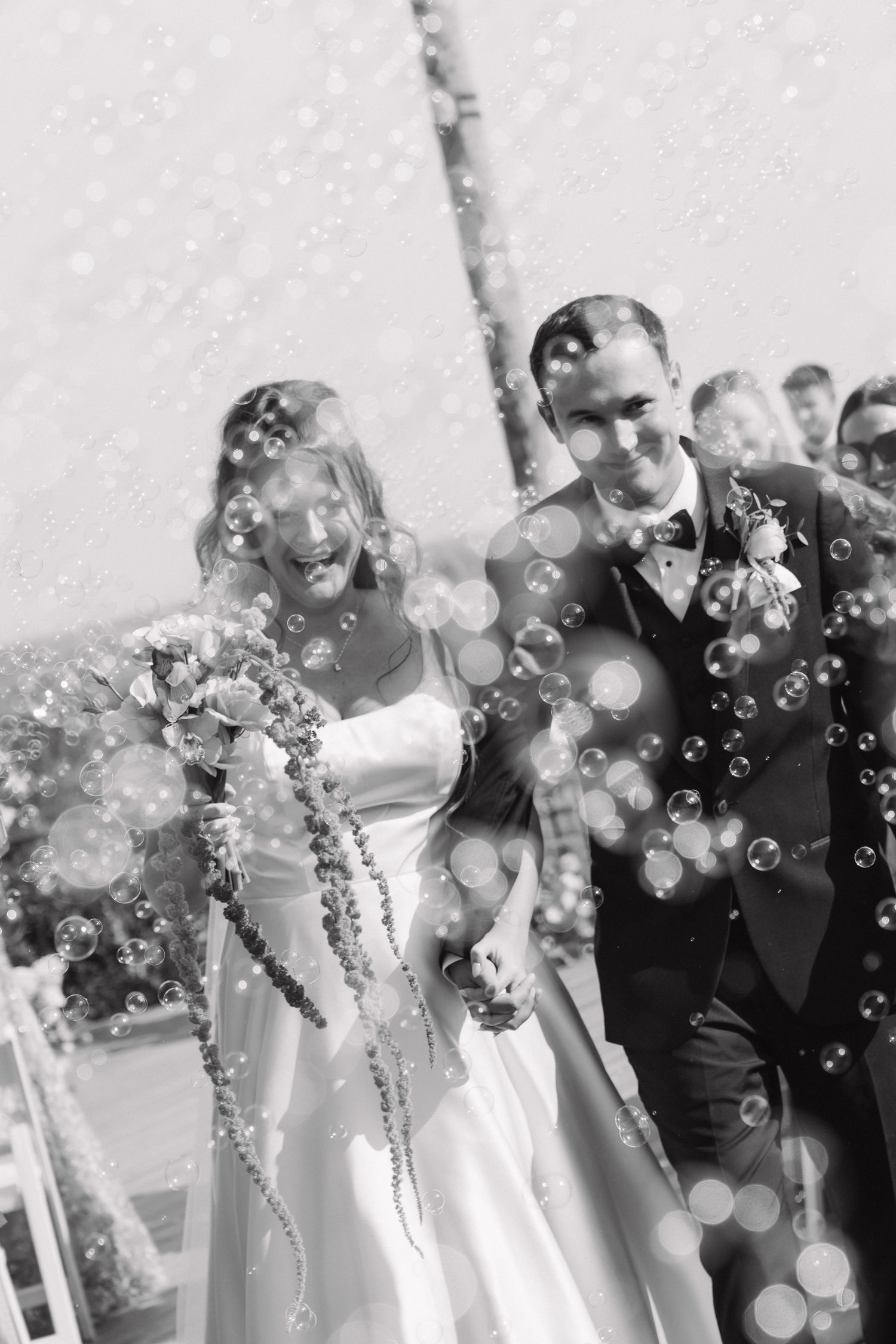 Black and white photo of a joyful bride and groom walking through bubbles after their wedding ceremony.