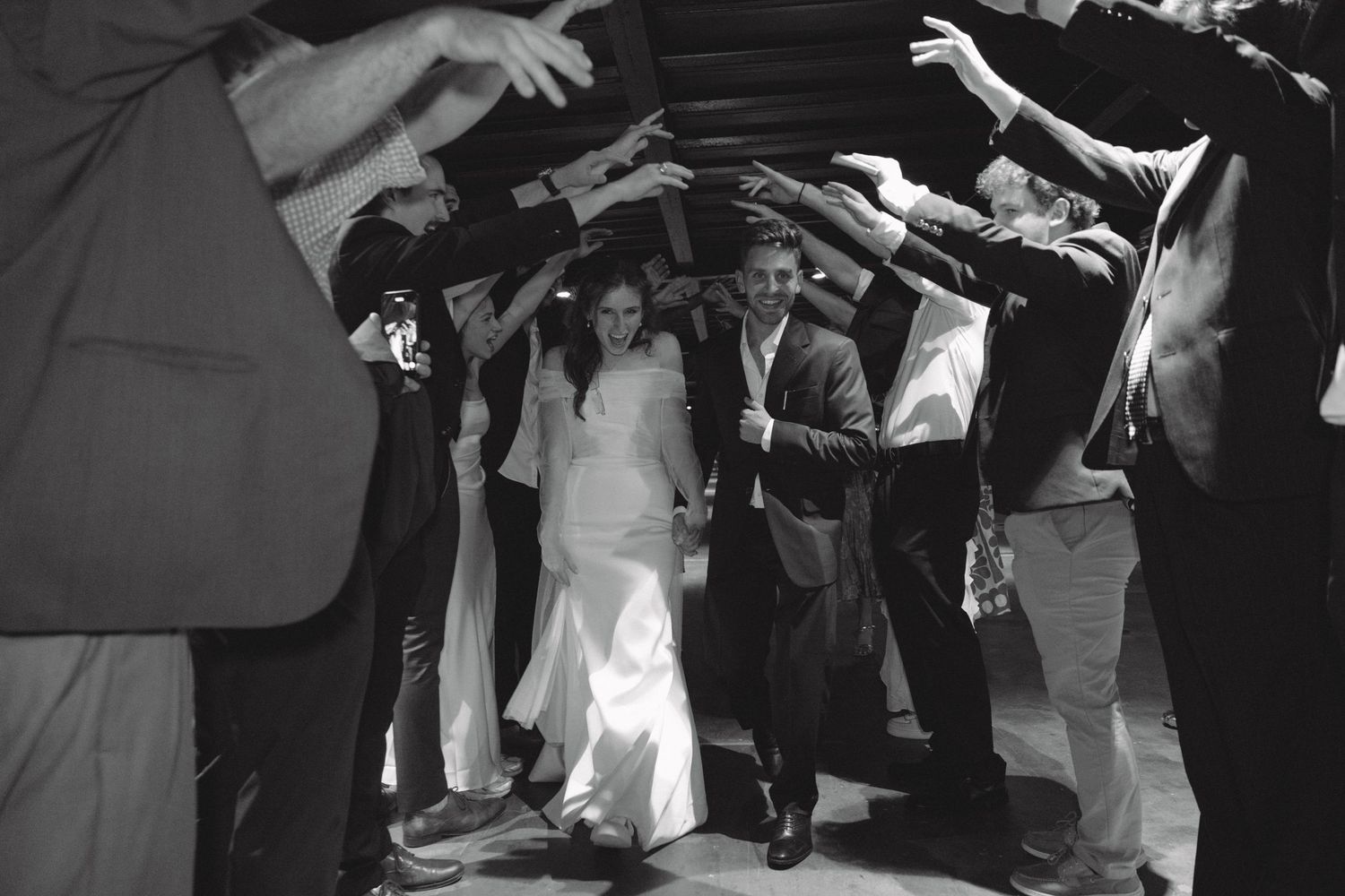 Black and white photo of newlyweds walking through a tunnel of raised hands at their wedding ceremony.