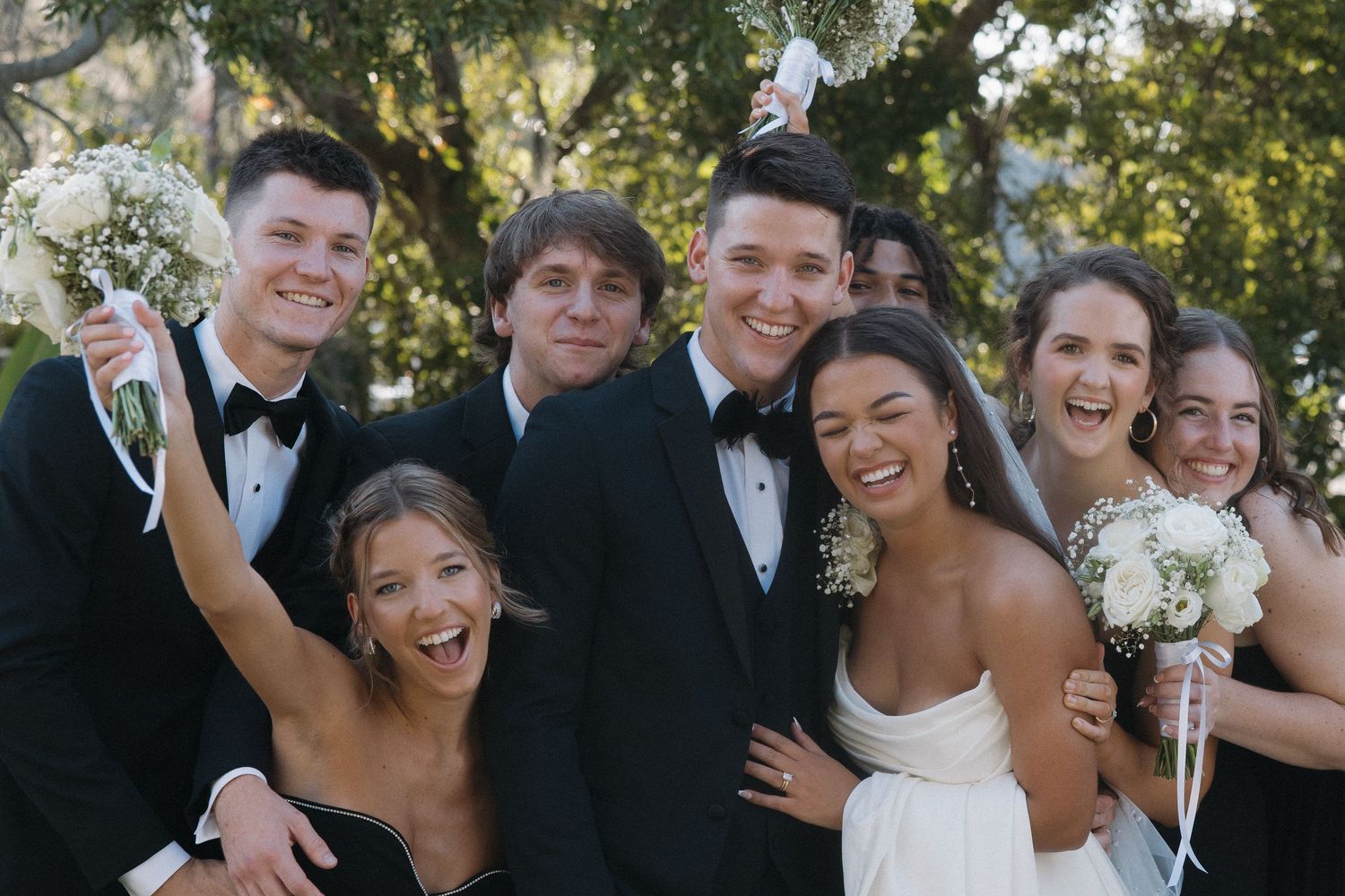 Group of young adults in formal attire smiling together outdoors, bridesmaids holding bouquets at a wedding.