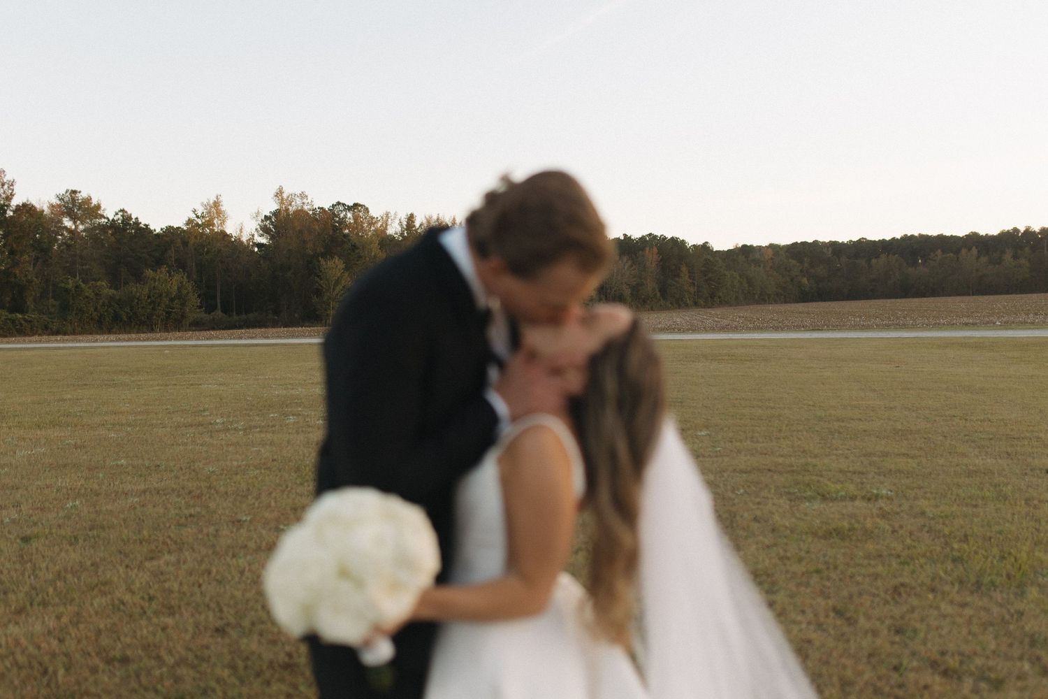 A bride and groom share a romantic kiss in an open field at golden hour, bride holding white bouquet.