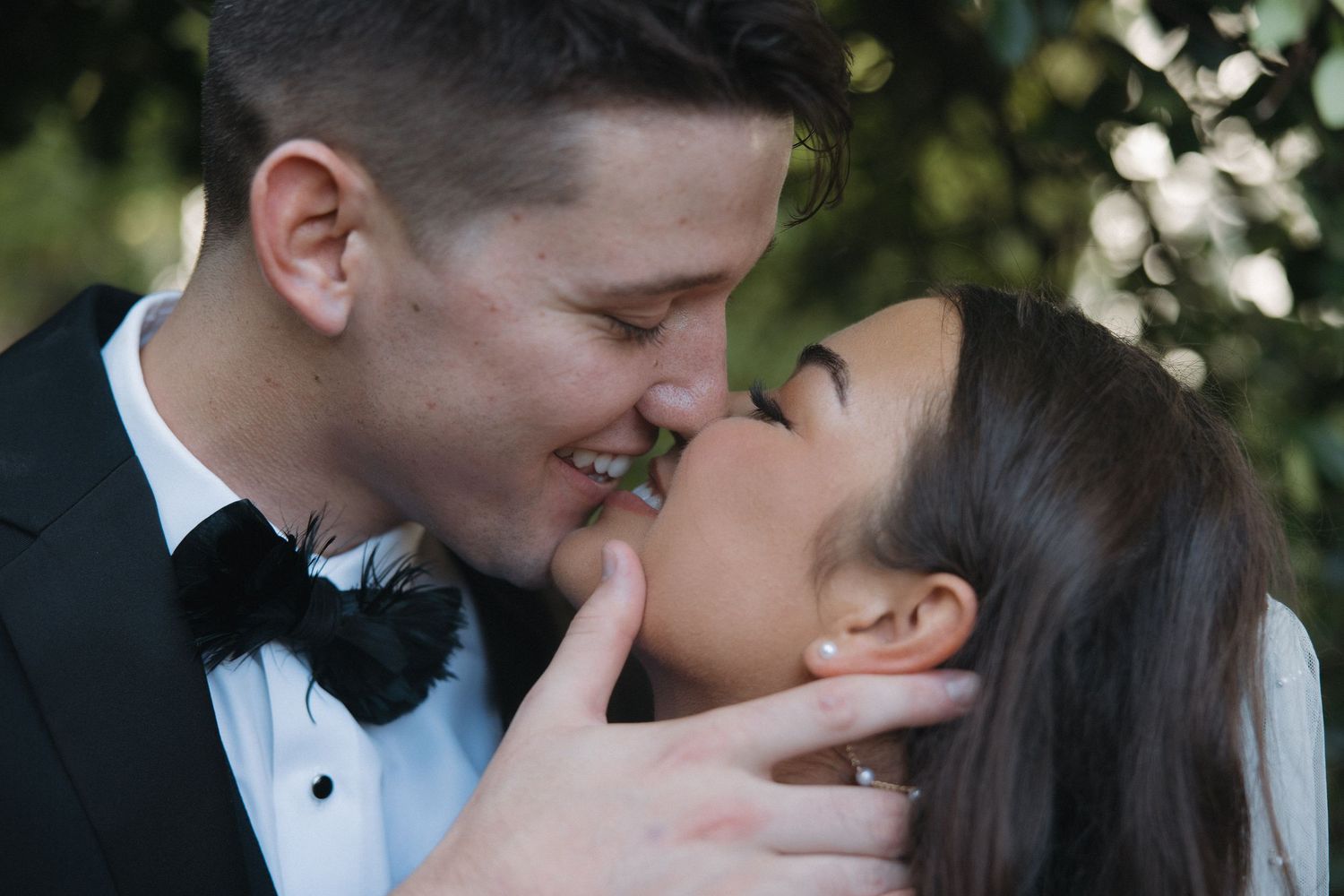 A couple in formal wedding attire shares a romantic almost-kiss, smiling closely together outdoors.