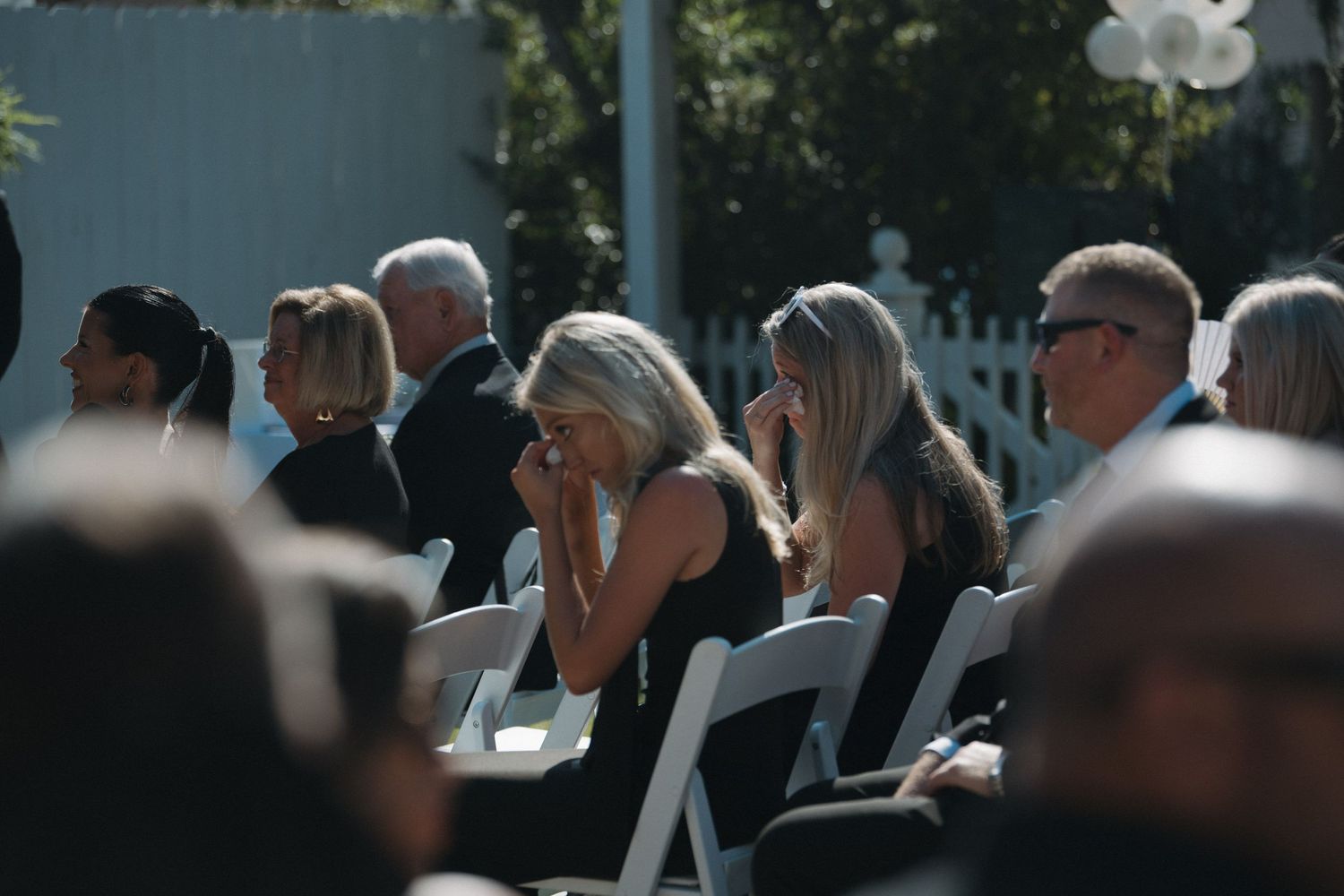 Emotional wedding guests dressed in black seated outdoors during a ceremony.