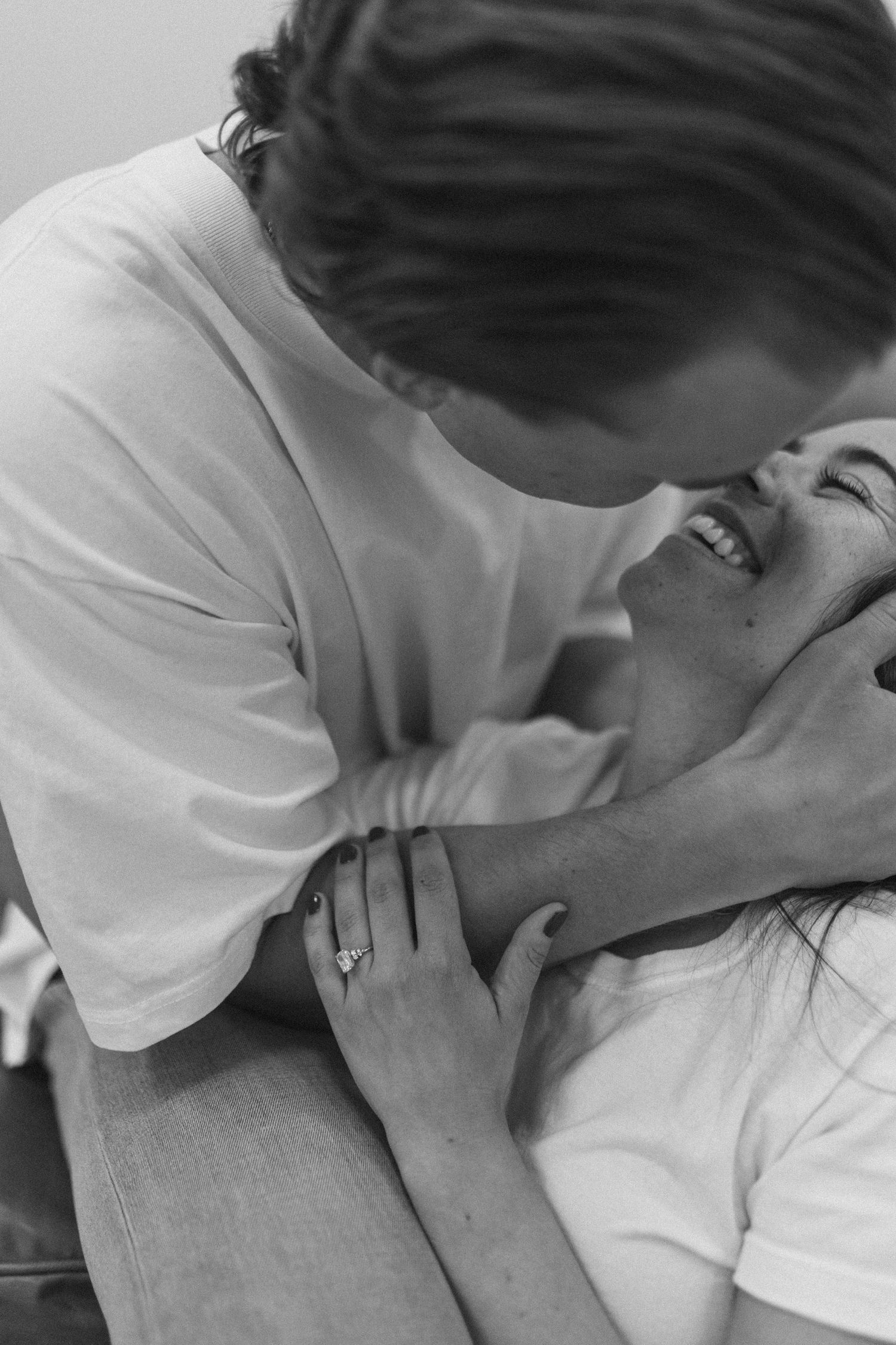 Black and white photo of couple laughing intimately, woman lying back with engagement ring visible on hand.