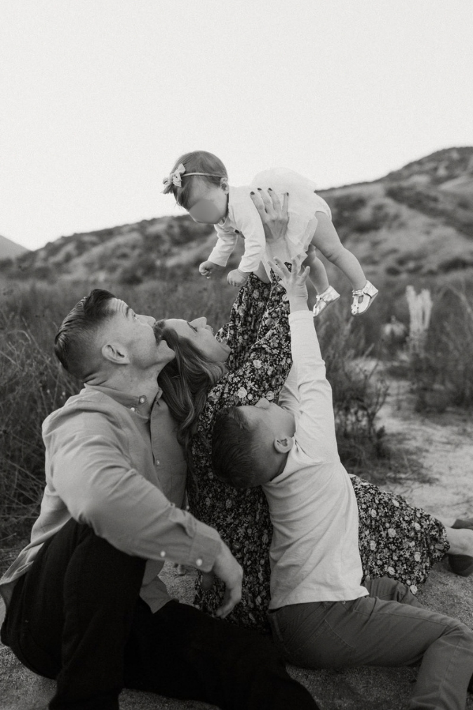Black and white photo of a couple sitting outdoors lifting a baby in the air, with hills and shrubs in the background.