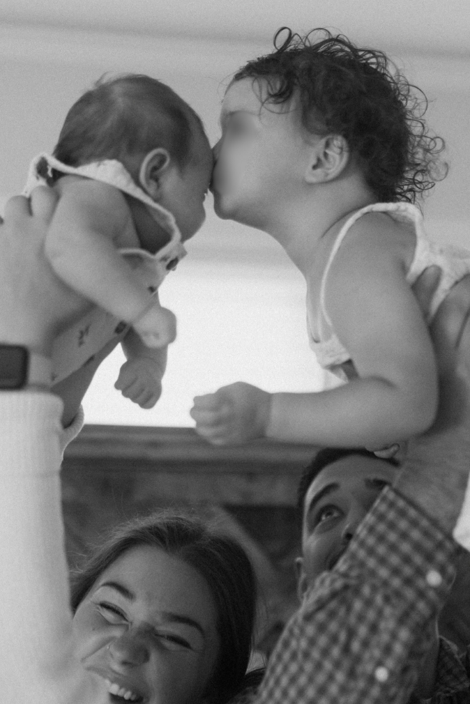 Black and white photo of a toddler kissing a newborn baby held up by parents smiling below.