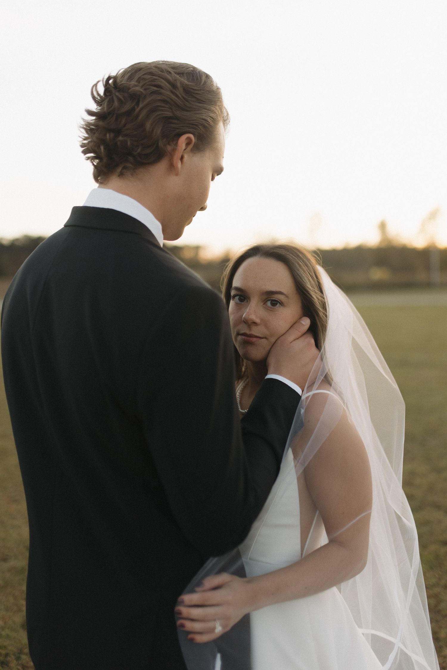 Bride and groom share a tender moment at sunset, with groom gently cupping bride's face as she gazes up lovingly.