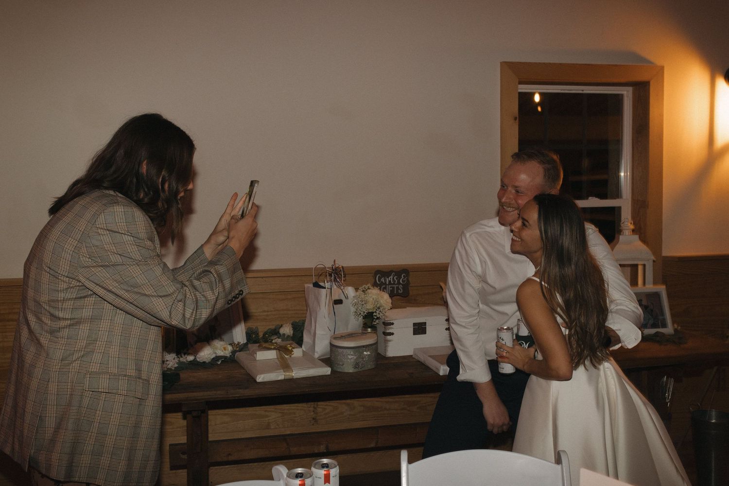 A wedding guest photographs a couple sharing a kiss at a rustic indoor reception venue.