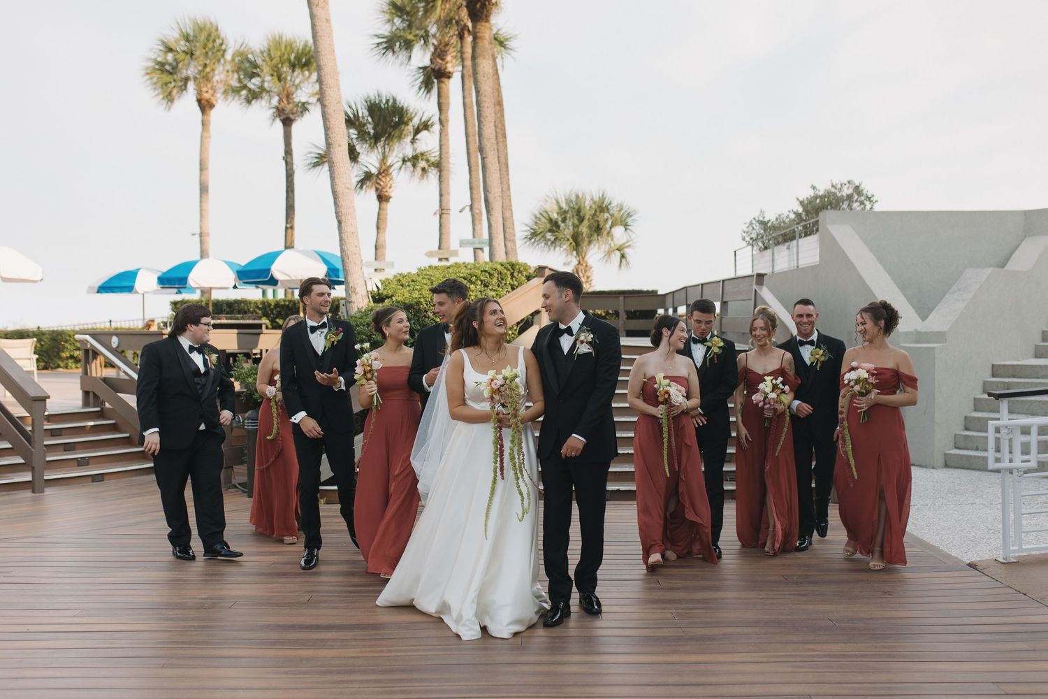 Wedding party in rust bridesmaid dresses and black tuxedos on a waterfront deck with palm trees.