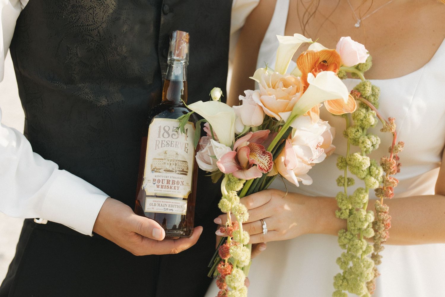 Bride and groom holding a vintage bourbon bottle and peach floral bouquet with green berry accents at wedding.