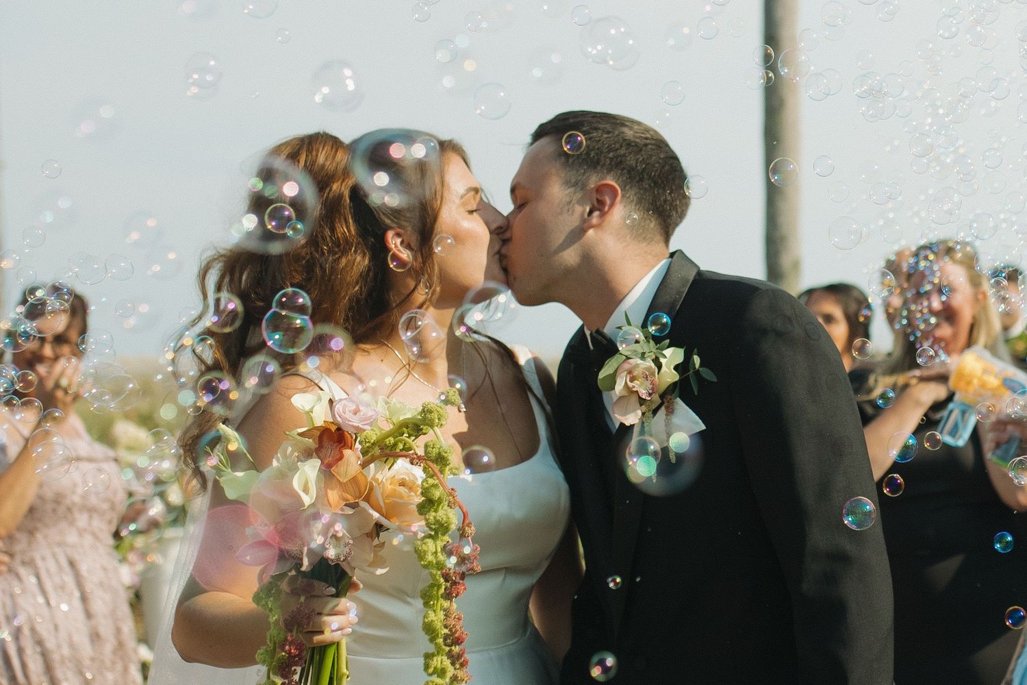 Newlyweds share a romantic kiss surrounded by bubbles, bride holding floral bouquet, guests celebrating in background.