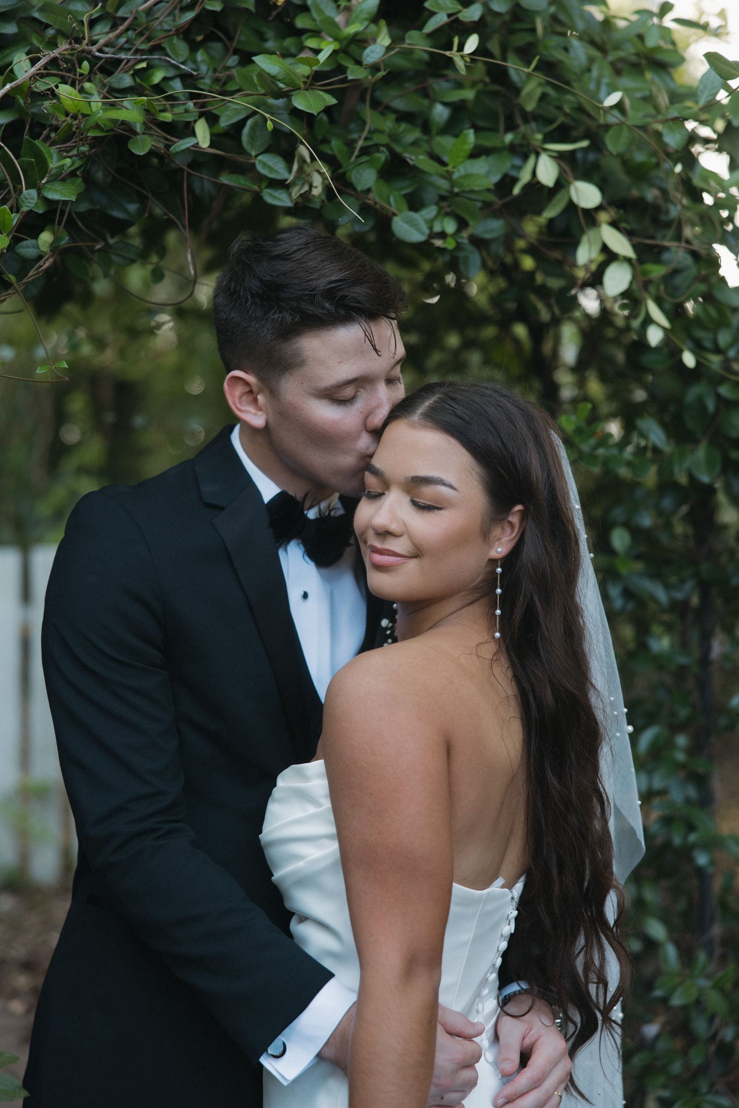Groom in black tuxedo kissing bride on cheek under green garden arch at outdoor wedding.