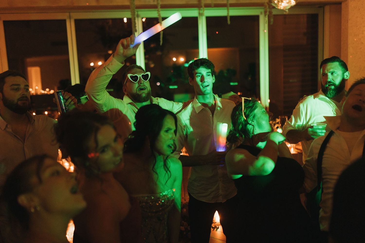 Group of young adults dancing at a dimly lit party with glowing neon light sticks and green ambient lighting.