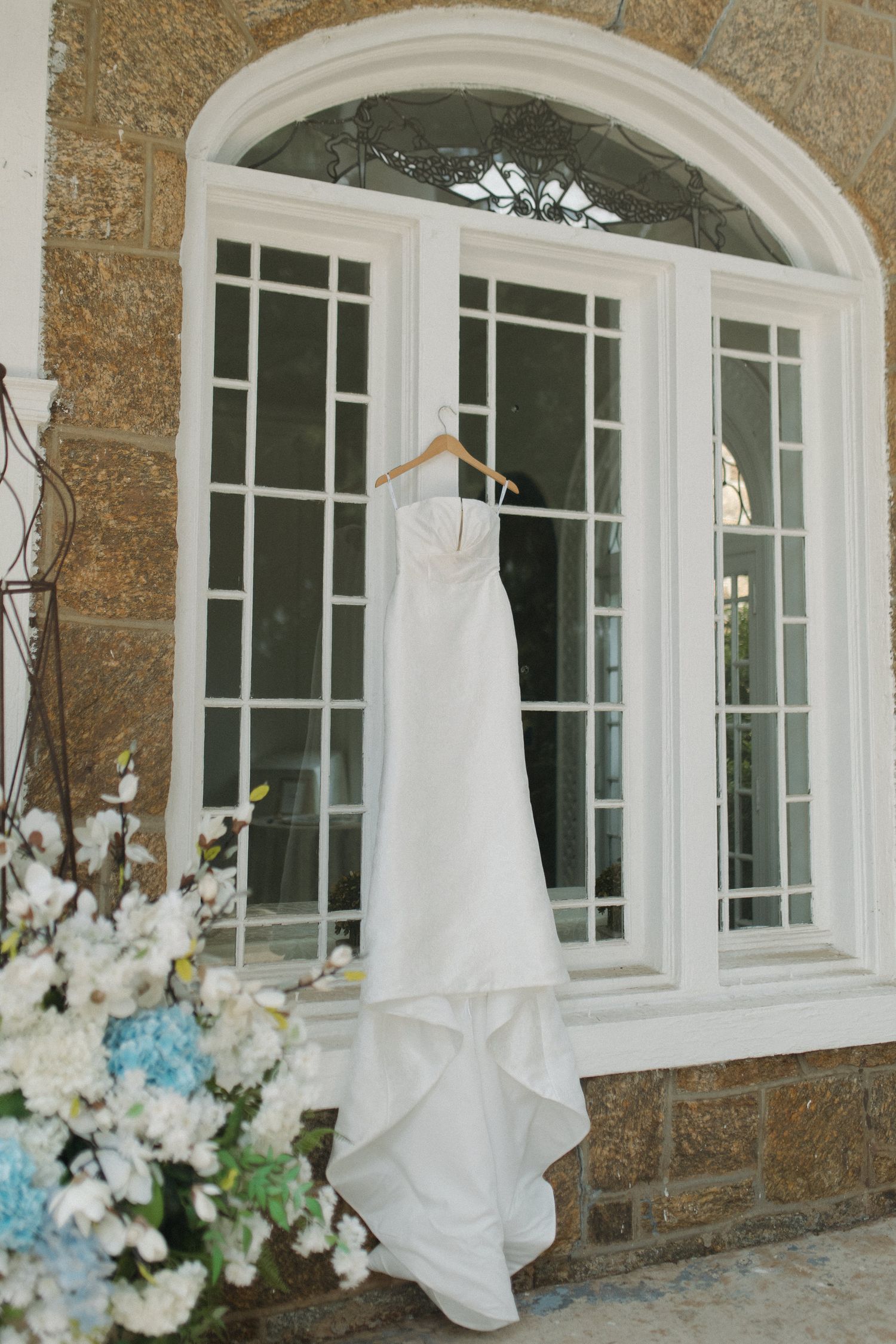White strapless wedding gown hanging on a hanger outside an arched window.