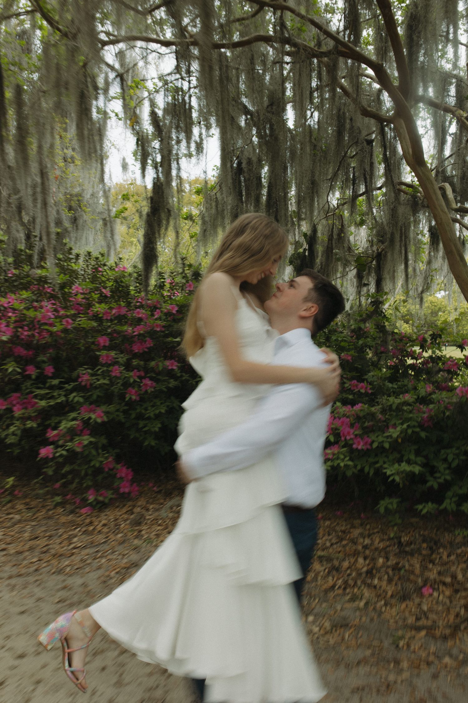 Groom lifting bride in white dress among pink azaleas and Spanish moss-draped trees in a romantic garden setting.