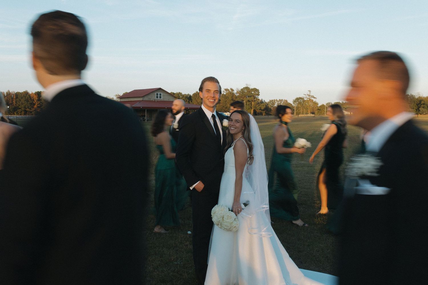 Bride and groom smiling outdoors at golden hour, surrounded by wedding guests in formal attire on a green lawn.
