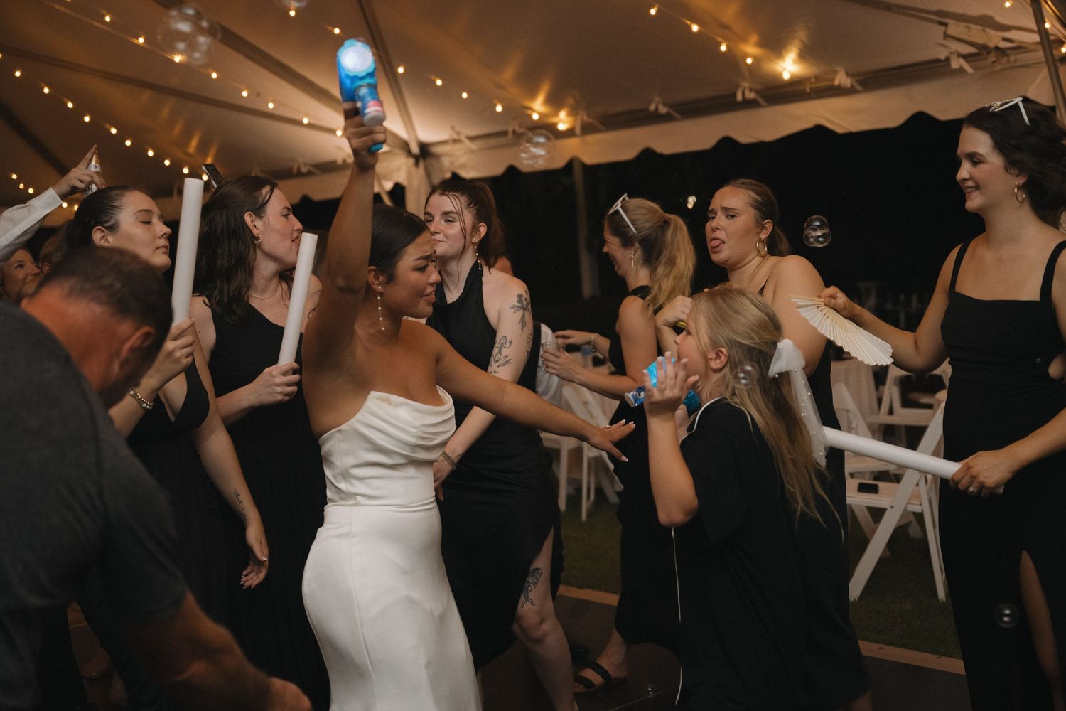 Bride in white gown dancing joyfully with guests holding glow sticks at wedding reception.