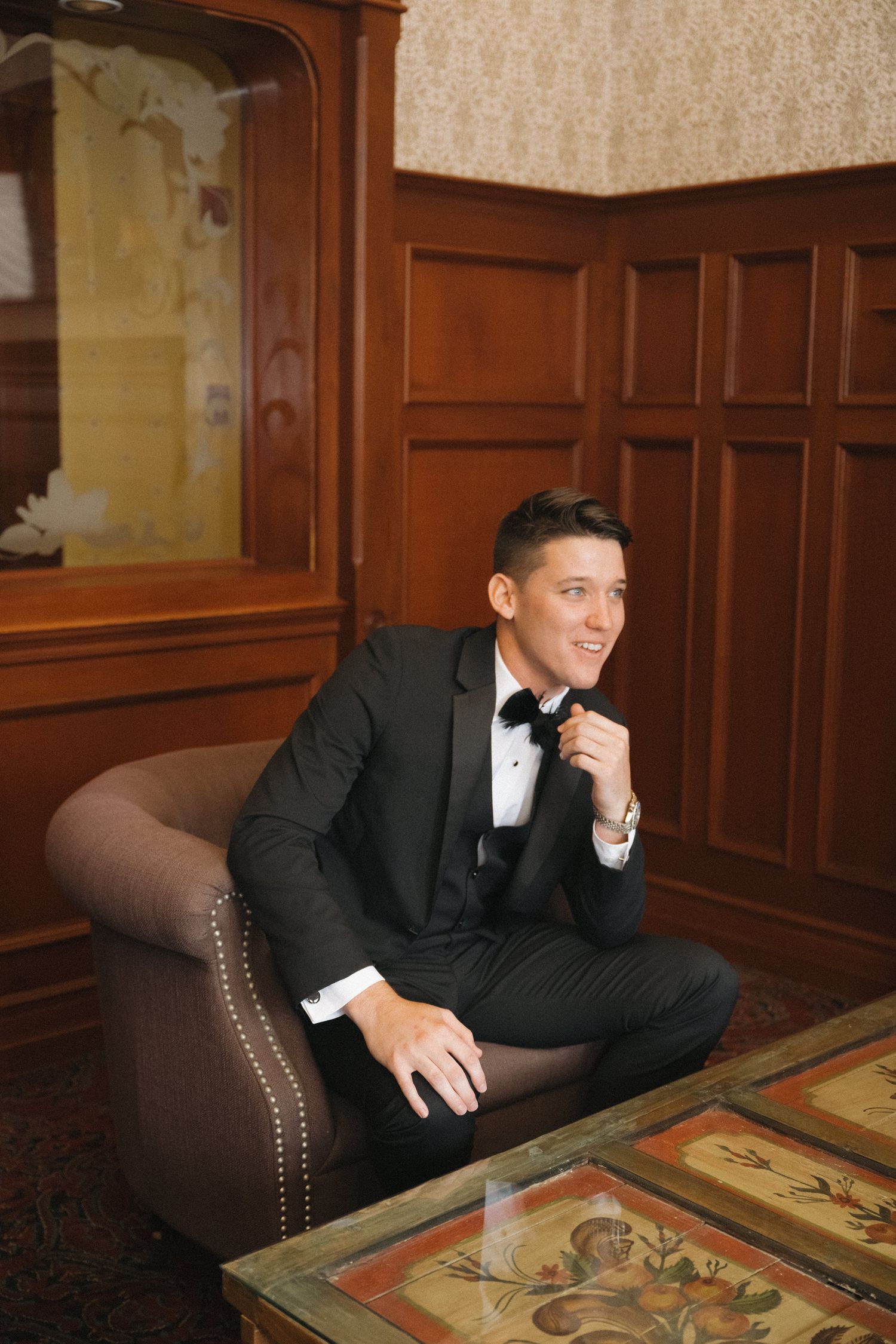 Groom in black tuxedo sitting in armchair adjusting bow tie in elegant wood-paneled room.