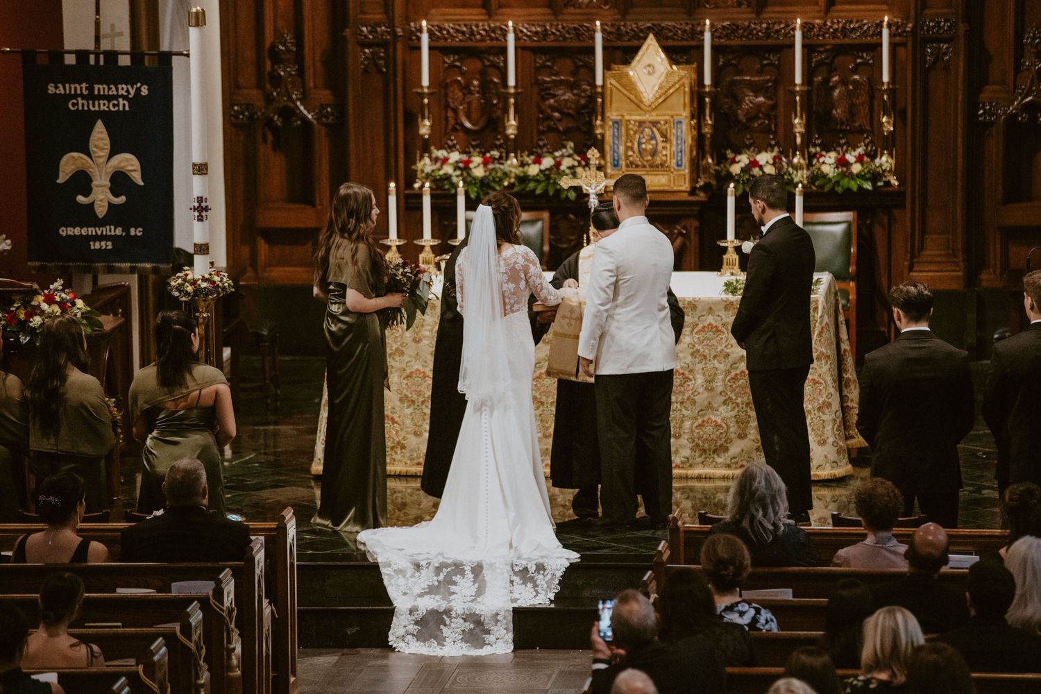 Bride and groom exchange vows at ornate church altar with floral arrangements and wedding party at Saint Mary's Church.