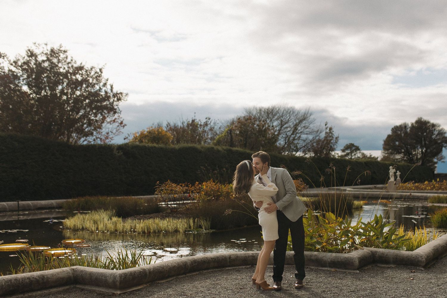 Couple shares romantic kiss in elegant garden setting with pond and hedges backdrop.