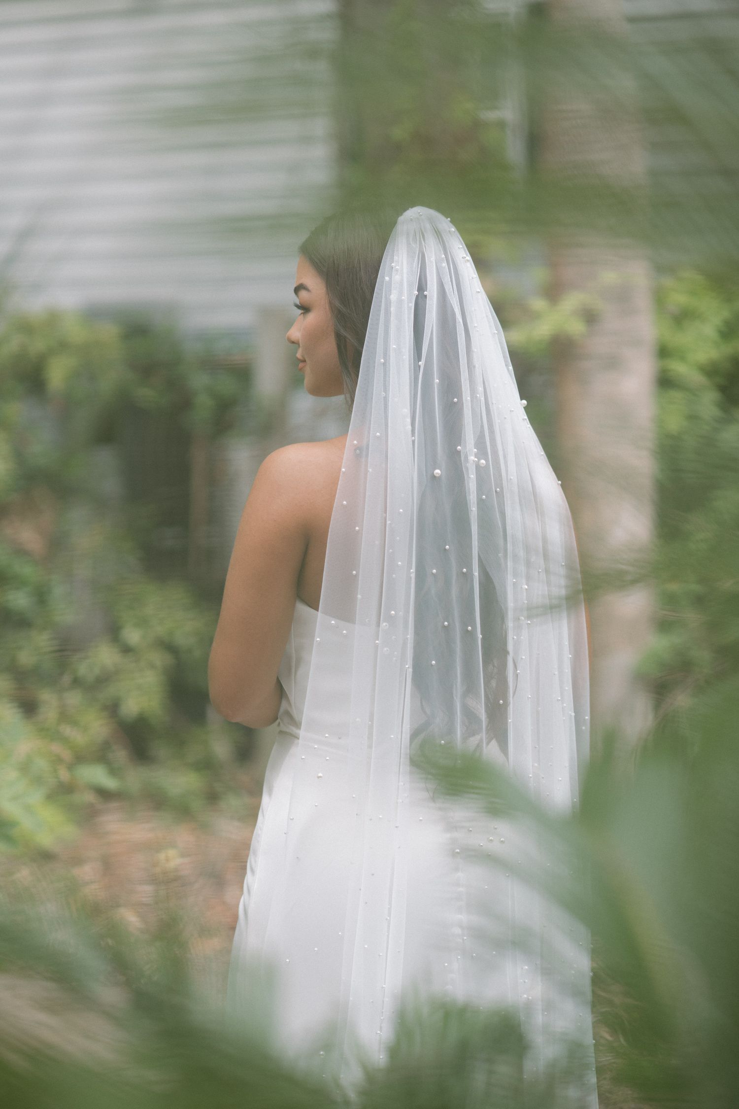 Elegant bride in white gown with pearl-embellished cathedral veil photographed from behind in lush garden setting.