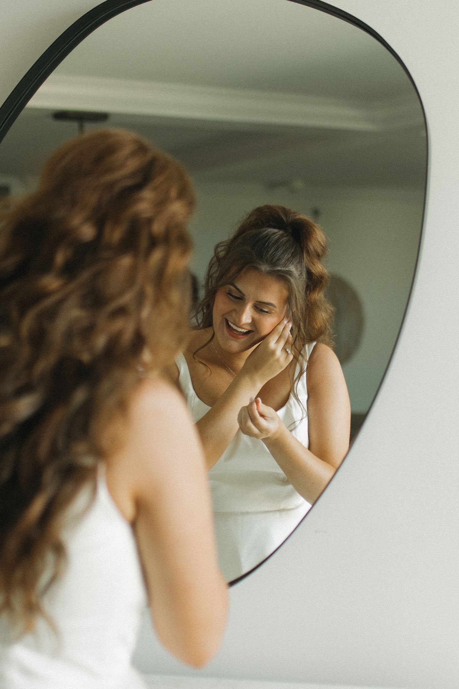 A smiling woman in a white dress applies skincare while looking at her reflection in a modern asymmetric mirror.
