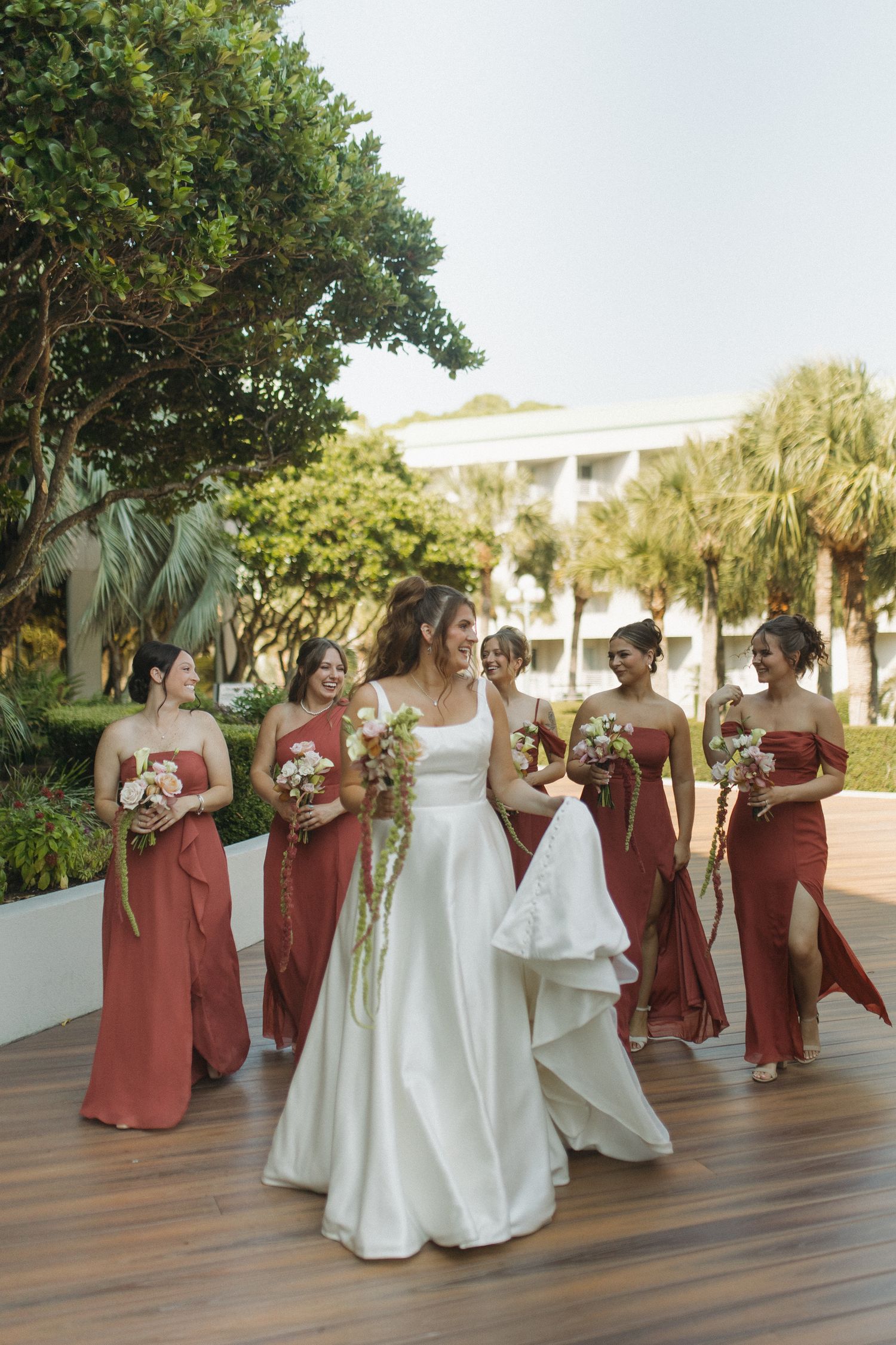 Bride in white gown walks with bridesmaids in rust-red dresses, holding bouquets on a wooden deck outdoors.