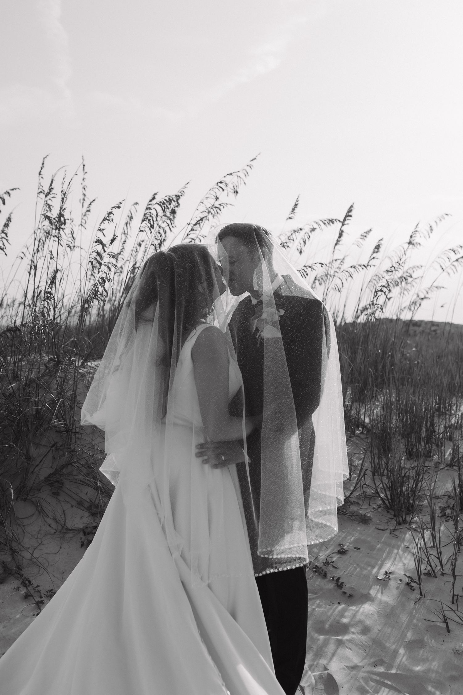 Black and white photo of bride and groom sharing a romantic moment under veil on sandy beach dunes.