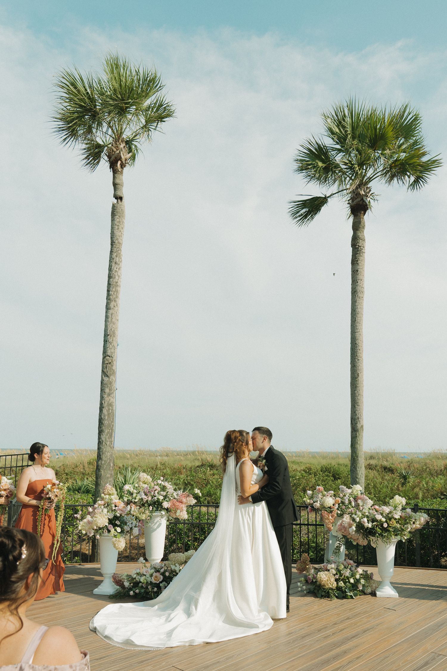 Bride and groom share first kiss at outdoor beach wedding ceremony flanked by tall palm trees.
