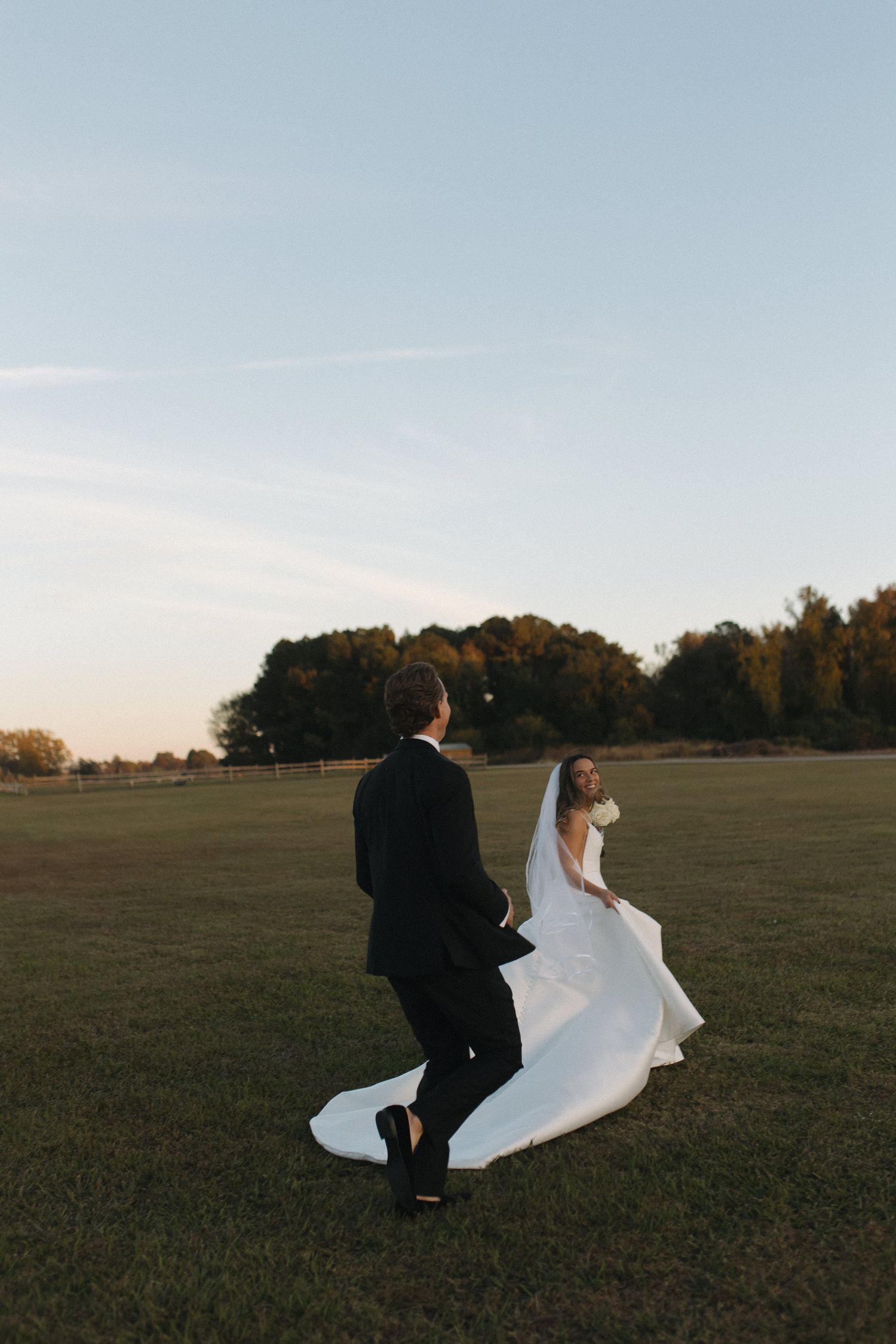 Bride in flowing white gown and groom in black suit walk across open field at sunset.