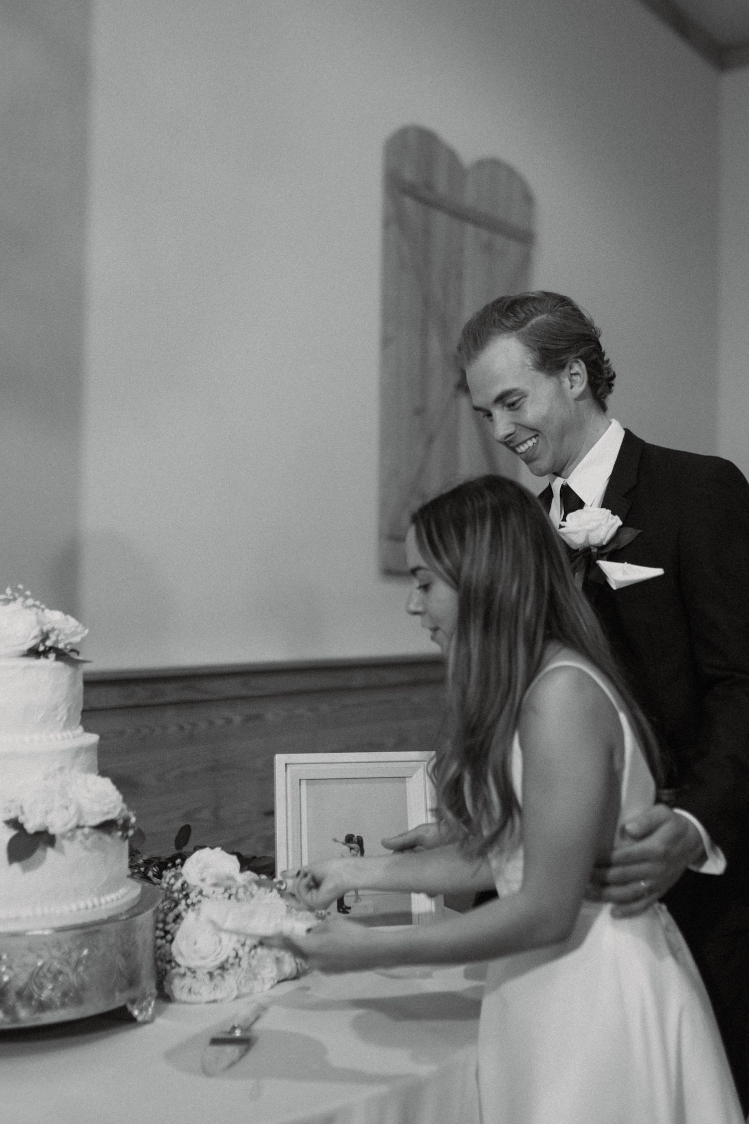 Black and white photo of bride and groom cutting wedding cake at elegant reception venue.