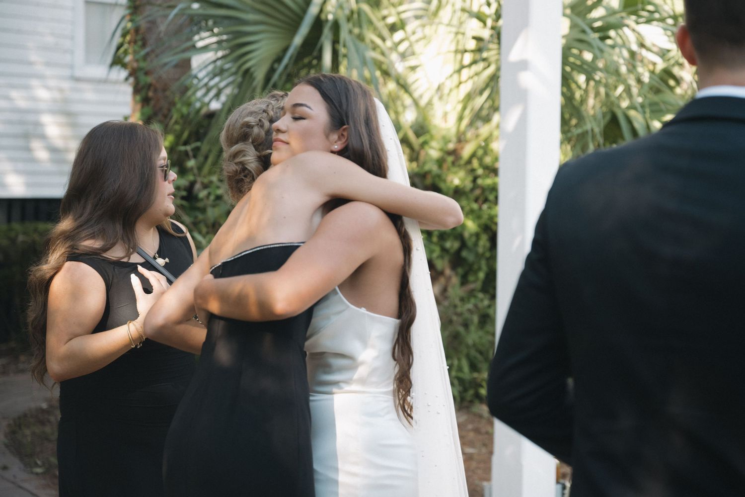Two women sharing a warm embrace at an outdoor wedding ceremony near a white pillar.