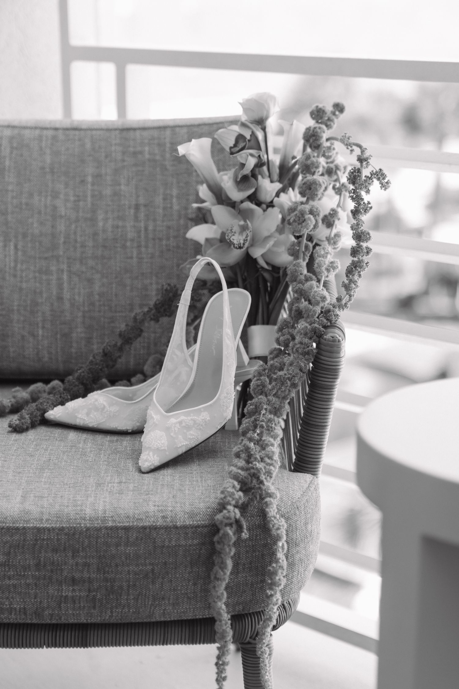 Black and white photo of elegant bridal heels beside a floral bouquet on a wicker chair.