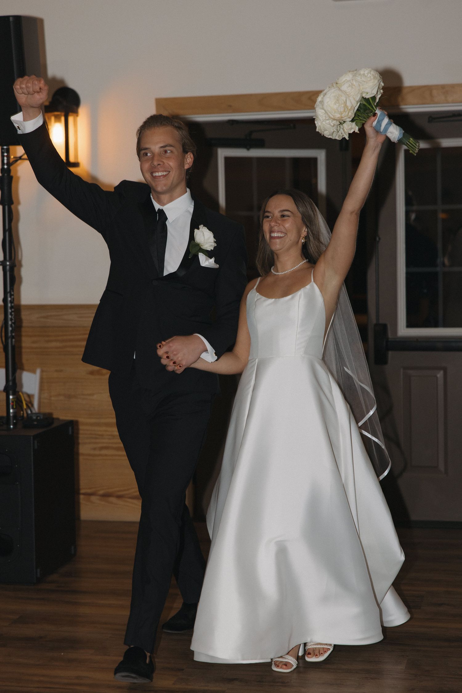 Joyful newlyweds celebrate their wedding reception entrance, bride in white gown holding bouquet, groom in black tuxedo.