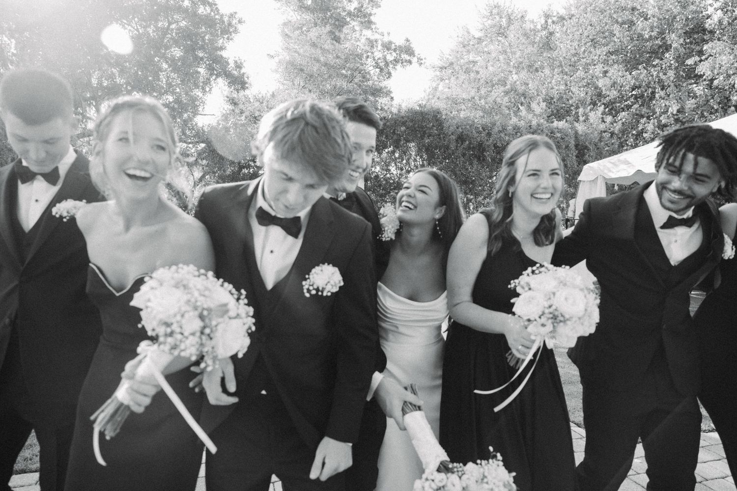 Black and white photo of a joyful wedding party in formal attire, laughing and celebrating outdoors.