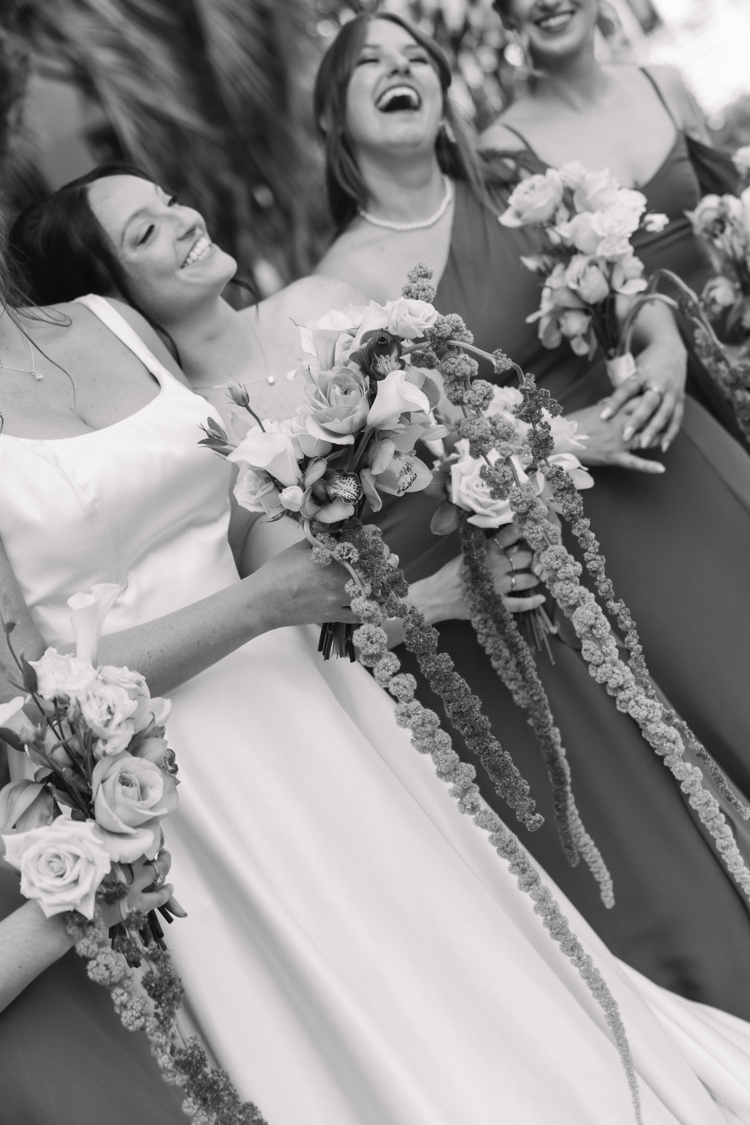 Black and white photo of a laughing bride holding a cascading floral bouquet with bridesmaids at a wedding.