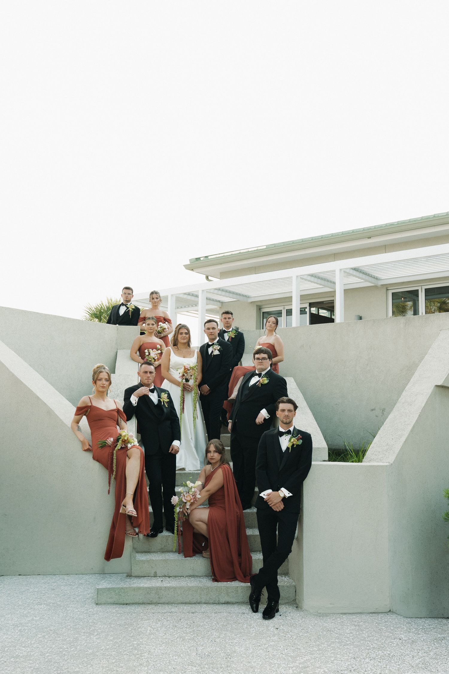 Wedding party poses on modern concrete steps, bridesmaids in rust dresses and groomsmen in black tuxedos.