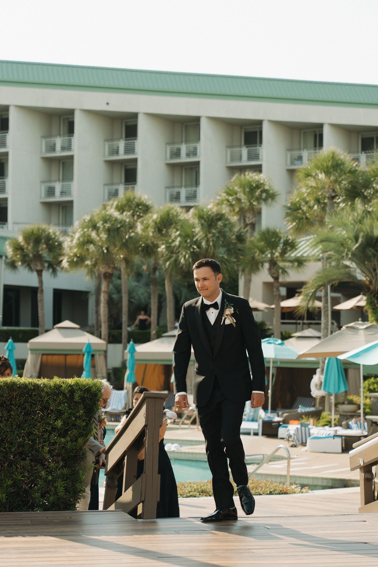 Groom in black tuxedo with boutonniere walks along poolside deck at tropical resort with palm trees.