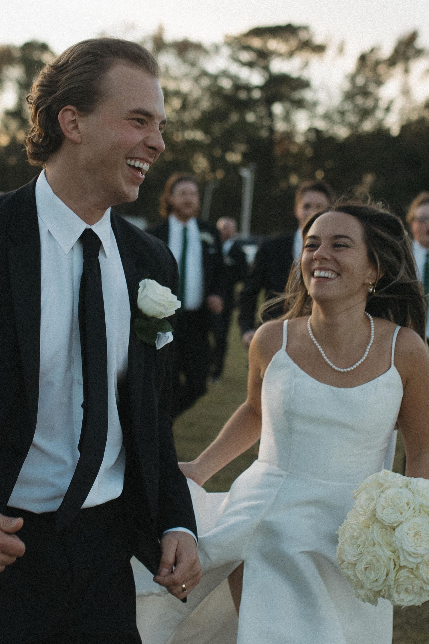 Smiling bride in white spaghetti strap gown and groom in black suit holding hands outdoors after wedding ceremony.