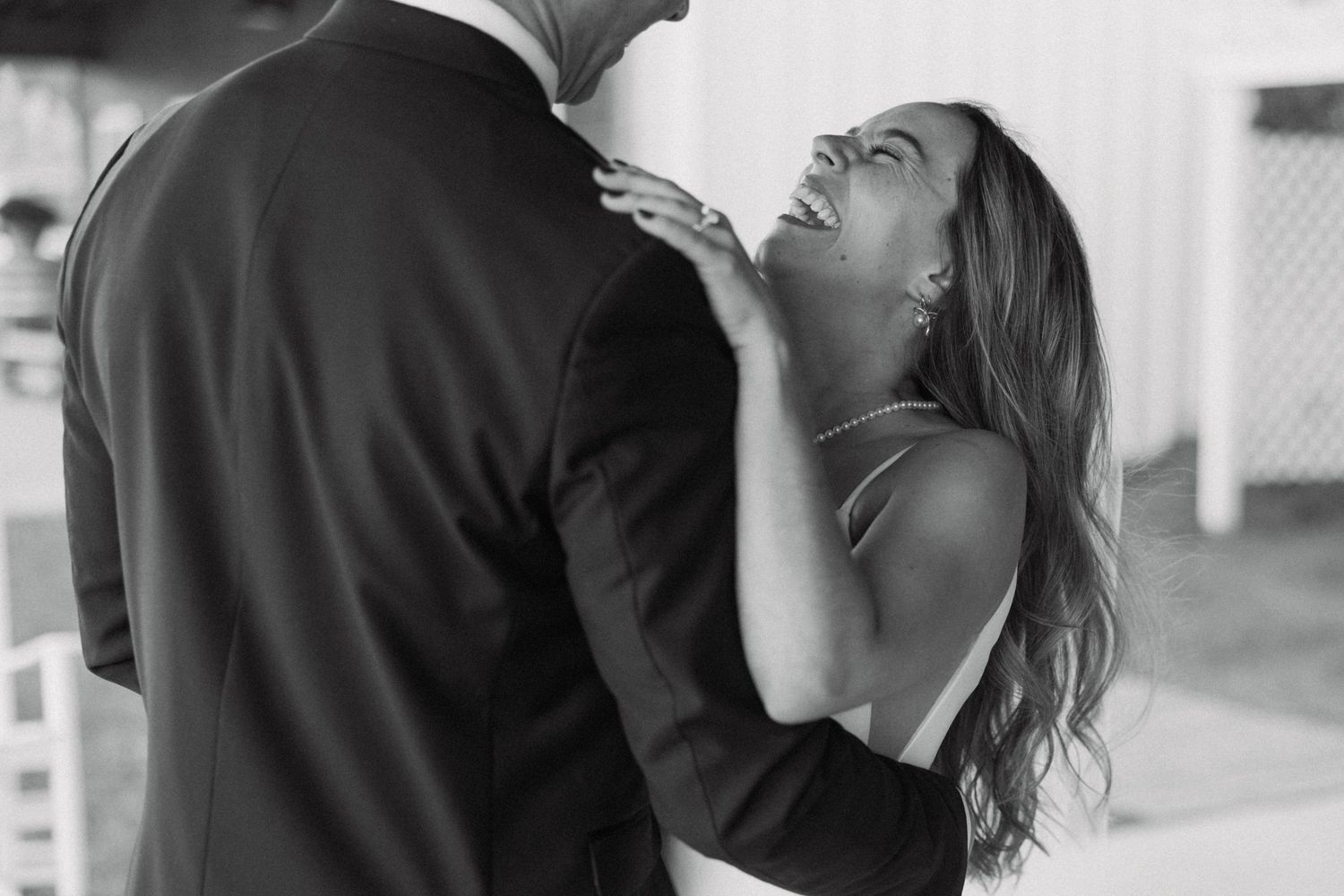 Black and white photo of a laughing bride embracing a groom in a suit during a romantic wedding moment.