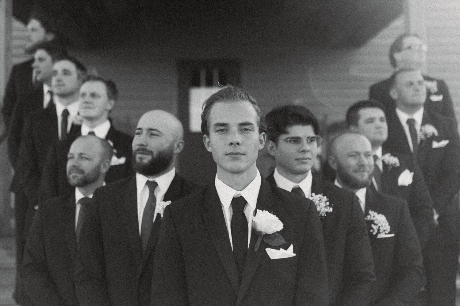 Black and white photo of groom and groomsmen in suits with boutonnieres standing outside a building.