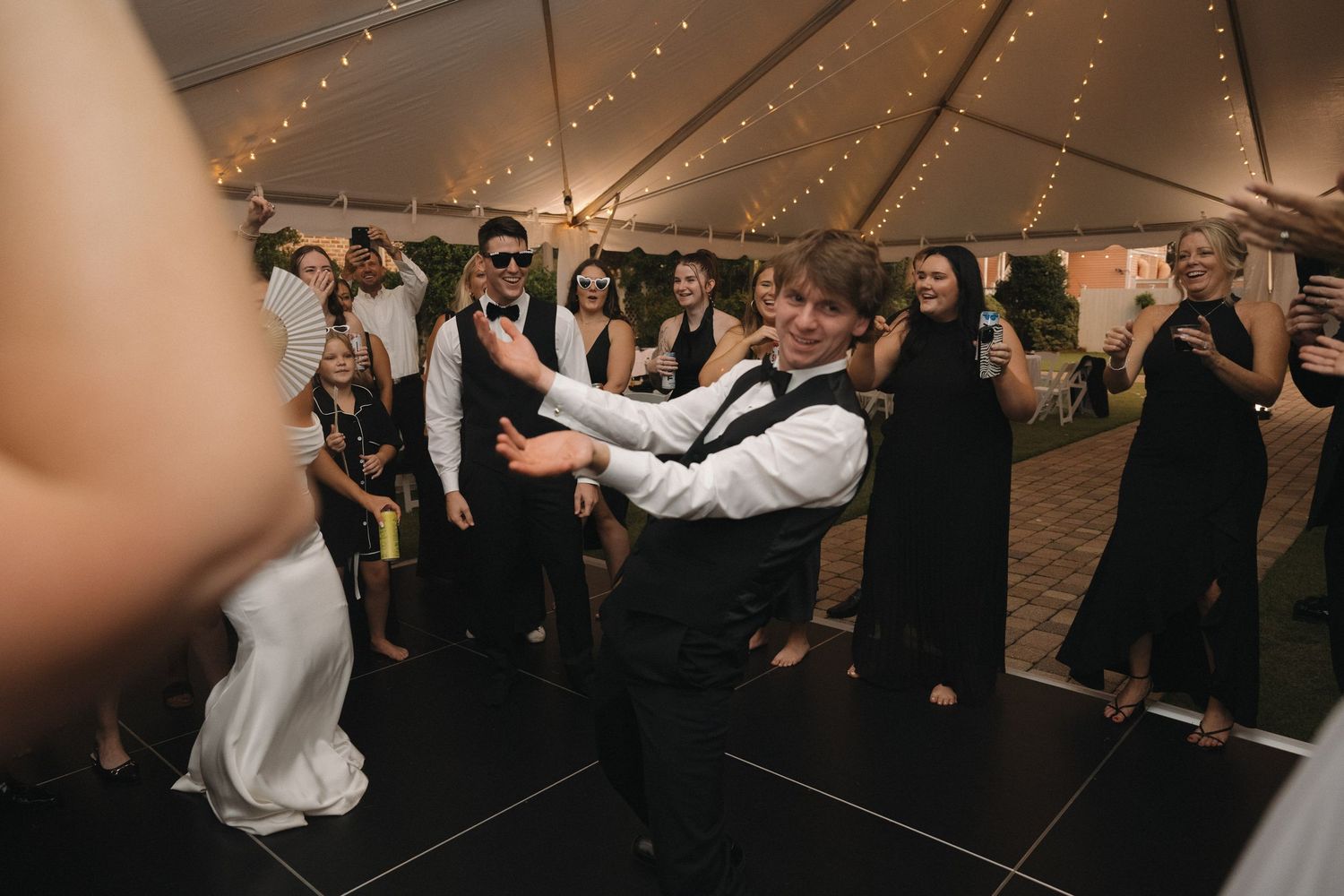 Wedding reception dance floor celebration under a tent with string lights, guests in formal attire dancing together.