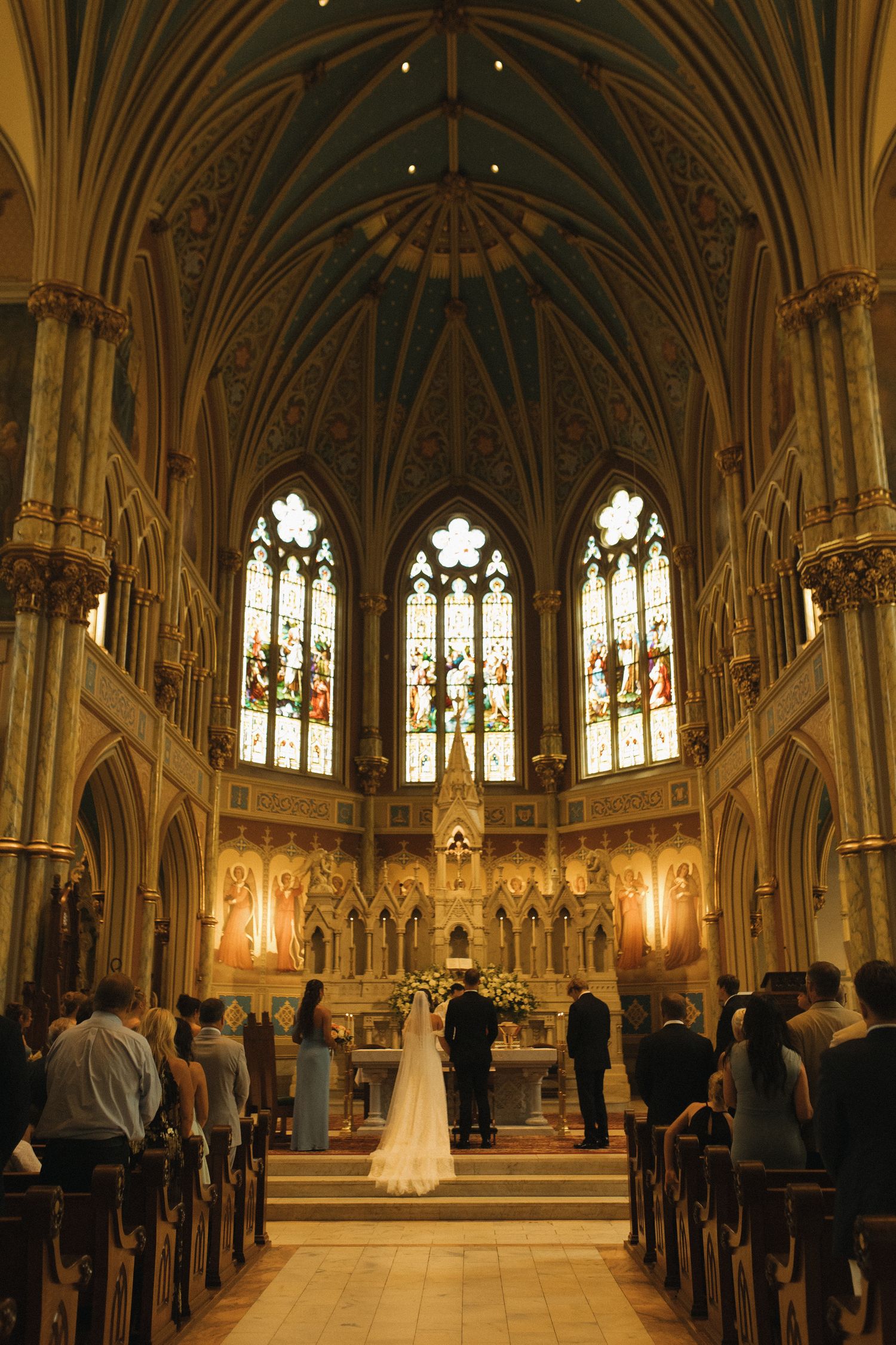 Elegant church wedding ceremony with bride and groom at ornate Gothic altar, stained glass windows, and wooden pews filled with guests.