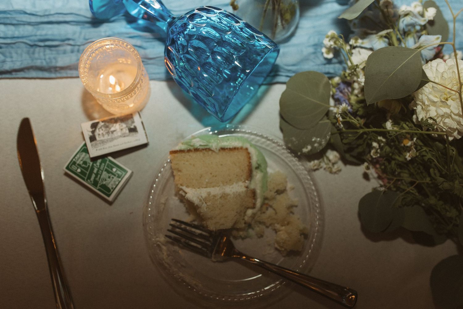 Slice of white wedding cake on glass plate with candle and floral decor.