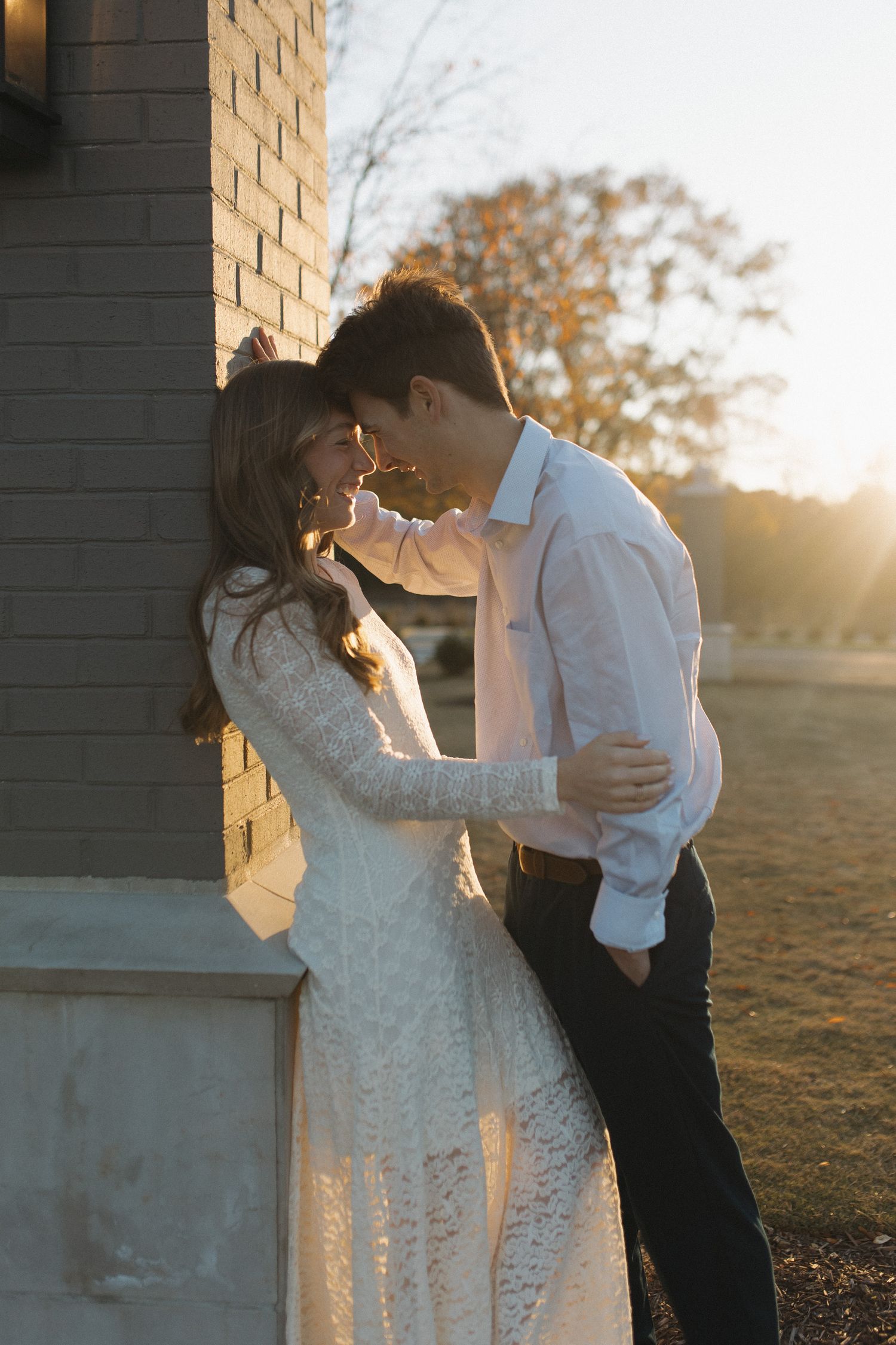 Couple sharing a tender kiss against a brick wall bathed in warm golden sunset light.