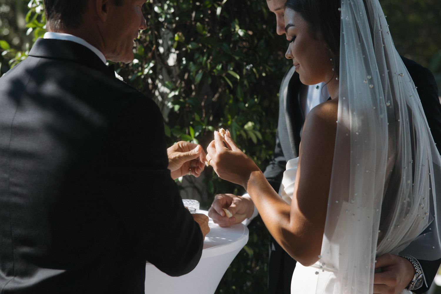 Bride and groom exchanging rings during an outdoor wedding ceremony, surrounded by lush green foliage.