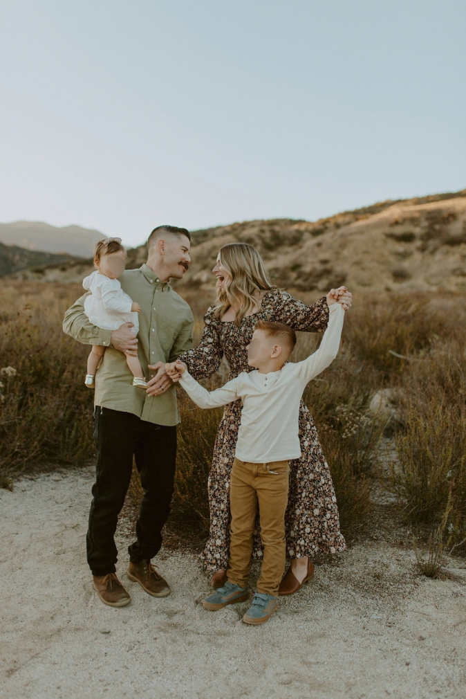 Family of four posing outdoors in golden hour light, parents holding children on a sandy desert trail with hills behind.