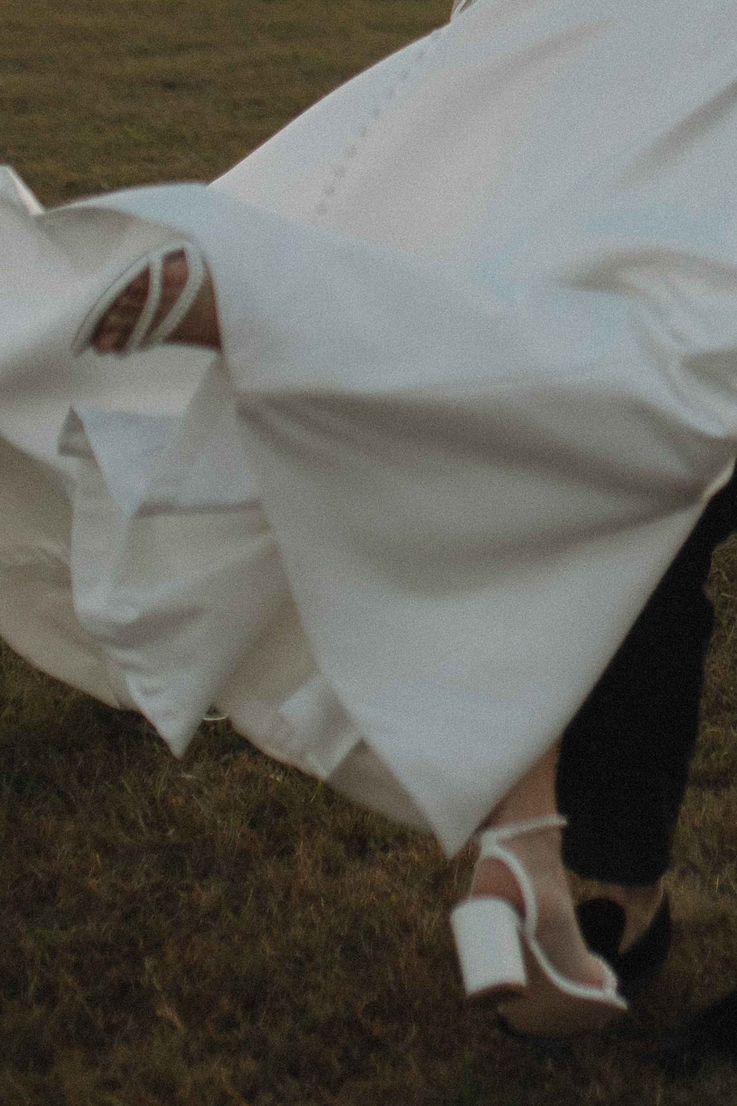 A flowing white wedding dress billows in the wind outdoors, revealing white heeled sandals on green grass.