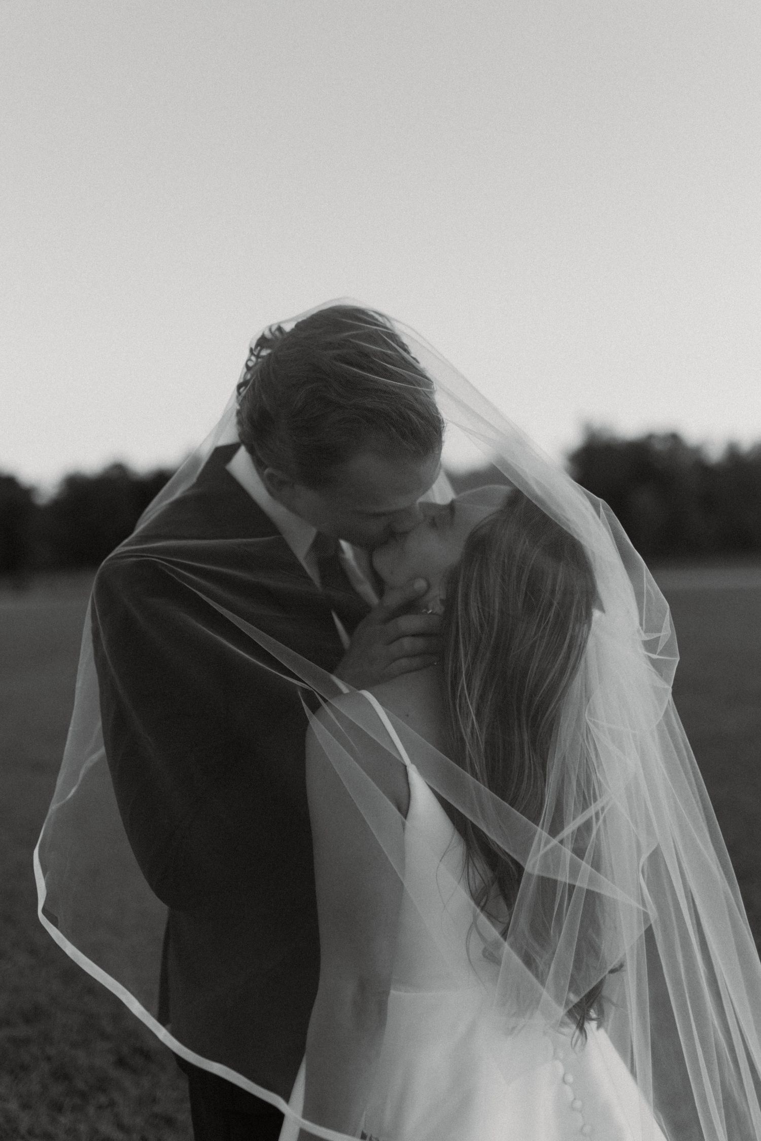 Black and white photo of a bride and groom sharing a kiss under a flowing veil in an outdoor field.